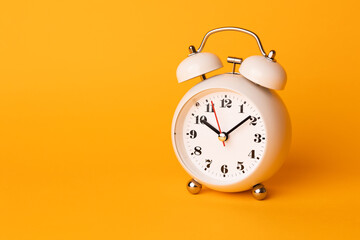 Small white alarm clock, black numbers, set the time placed on a table. Clock on isolated yellow background.