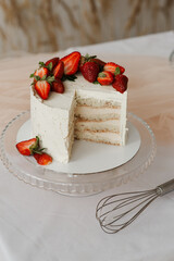 white cream cake with strawberries on a tray and whisk on a white table