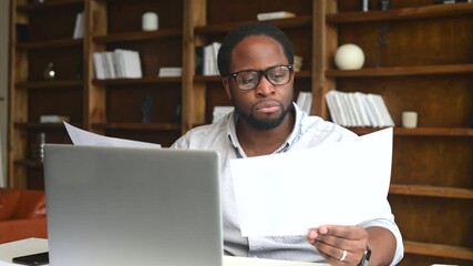Clever and concentrated African-American guy is doing paperwork, a man looking through workpaper sitting at the desk with a laptop in office, a man checking report, collate data on the laptop - Powered by Adobe