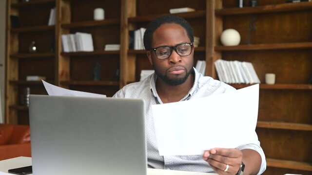 Clever and concentrated African-American guy is doing paperwork, a man looking through workpaper sitting at the desk with a laptop in office, a man checking report, collate data on the laptop
