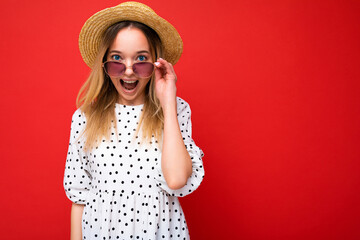 Attractive amazed young blonde woman wearing everyday stylish clothes and modern sunglasses isolated on colorful background wall looking at camera with open mouth