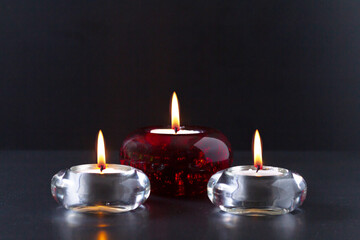 Mourning or prayer concept. Three burning candles on black background. Two of them in transparent glass candlesticks and one in red glass chandelier.