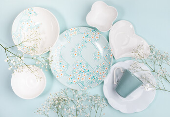 Spring festive layout of beautiful dishware white and with floral decor and white flowers on light blue. Top view.