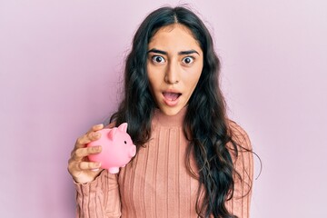 Hispanic teenager girl with dental braces holding piggy bank scared and amazed with open mouth for surprise, disbelief face