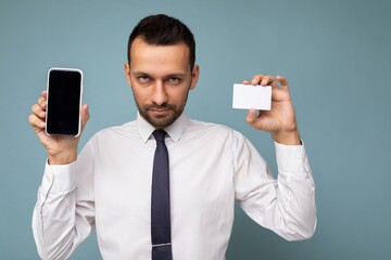 Closeup photo of handsome good looking brunet man with beard wearing casual white shirt and tie isolated on blue background with empty space holding in hand and showing mobile phone with empty screen