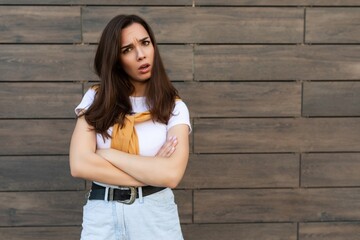 Portrait of sad upset sorrowful dissatisfied and asking young brunet woman wearing casual white t-shirt with yellow sweater poising near brown wall in the street