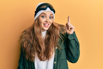Beautiful blonde caucasian woman wearing snow wear and sky glasses pointing finger up with successful idea. exited and happy. number one.