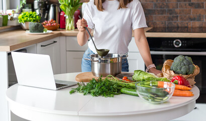 Close up woman in white t-shirt cooking soup with fresh vegetables in kitchen at home. Menu, recipe...