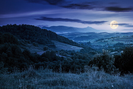mountainous rural landscape at night. beautiful scenery with forests, hills and meadows in full moon light. ridge with high peak in the distance. village in the distant valley