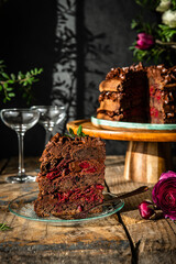 Piece of homemade chocolate and cherry cake, whole cake  on wooden stand, glasses and flowers on dark background.