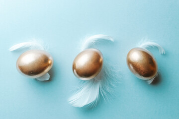 Golden eggs, white feathers on pastel blue background in Happy Easter decoration. Foil minimalist egg design, modern top view design.