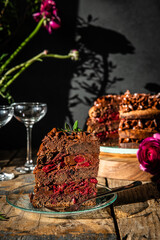 Piece of homemade chocolate and cherry cake, whole cake  on wooden stand, glasses and flowers on dark background.