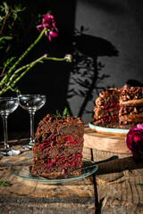 Piece of homemade chocolate and cherry cake, whole cake  on wooden stand, glasses and flowers on dark background.