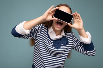 Attractive positive amazed brunette girl wearing striped longsleeve standing isolated on blue background with copy space holding smartphone showing phone in hand with empty screen display for mockup