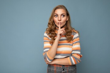 Shot of young blonde wavy-haired woman with sincere emotions wearing striped sweater isolated on blue background with copy space and showing shhh gesture. Keep secret
