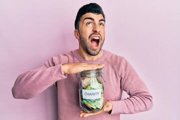 Young hispanic man holding charity jar with money angry and mad screaming frustrated and furious, shouting with anger looking up.