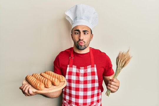 Young hispanic man wearing baker uniform holding homemade bread and spike wheat making fish face with mouth and squinting eyes, crazy and comical.