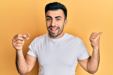 Young hispanic man holding deafness headset pointing thumb up to the side smiling happy with open mouth