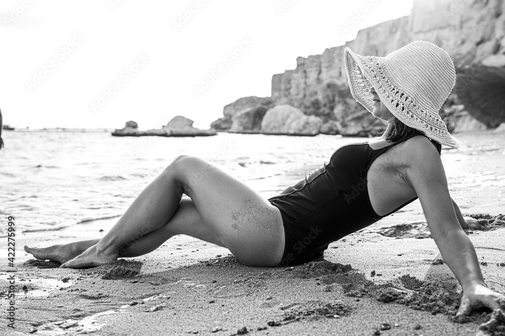 Wall mural Close up of a young woman in a hat and swimsuit lies on the sand on the seashore.