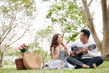 Fototapeta premium Handsome young Vietnamese man playing guitar and singing song for girlfriend when they are having picnic in park
