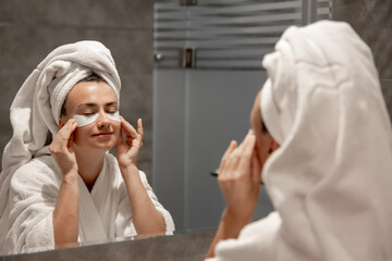 A young woman in a robe and with a towel on her head is putting patches under her eyes.