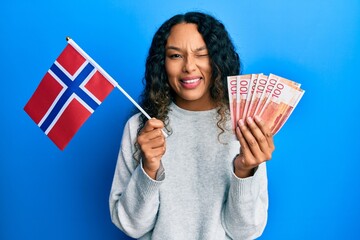 Young latin woman holding norway flag and norwegian krone banknotes winking looking at the camera...