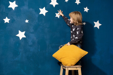 Little child boy in pyjamas with pillow playing, dreaming of space, decorate bedroom wall by white stars. Caucasian kid on stool on blue background with blank space for children store advertising