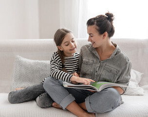 Young mother and daughter have fun together, reading a book at home on the couch.