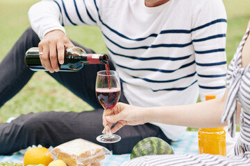 Cropped image of young man pouring red wine in glass of girlfriend when they are enjoying picnic in park