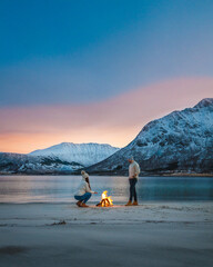 A couple enjoying the fire at the beach in Norway during the Blue hour,  enjoying a sunset with a beautiful colourful sky and majestic mountains behind them.