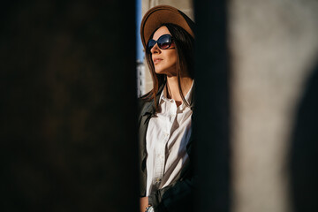 Outdoor fashion portrait of elegant, luxury woman wearing beige hat, sunglasses, trendy white shirt, in a green trench coat, walking in street of European city. Copy, empty space for text