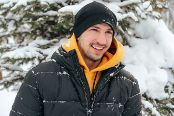 Fototapeta na wymiar Young man in hoodie and warm jacket standing in a snowy park and smiling