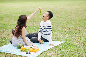Pretty young woman feeding boyfriend with grapes when they are sitting on blanket in city park