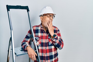 Handsome mature handyman close to construction stairs wearing hardhat bored yawning tired covering...