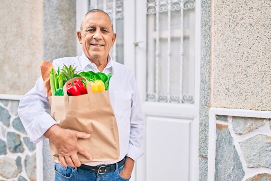Senior man smiling happy holding paper bag with food walking at the city.