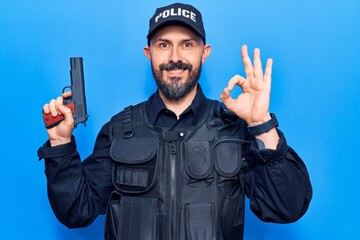 Young handsome man wearing police uniform holding gun doing ok sign with fingers, smiling friendly gesturing excellent symbol