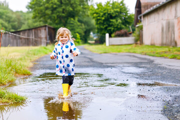 Naklejka premium Little toddler girl wearing yellow rain boots, running and walking during sleet on rainy cloudy day. Cute happy child in colorful clothes jumping into puddle, splashing with water, outdoor activity