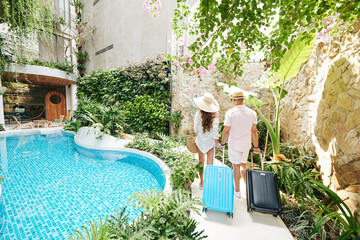 Young couple with suitcases walking to room of luxurious spa resort, view from the back