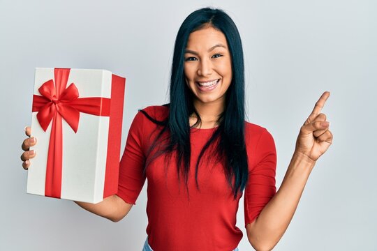 Beautiful hispanic woman holding gift smiling happy pointing with hand and finger to the side