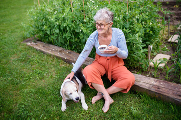 Portrait of senior woman with coffee sitting outdoors in garden, pet dog friendship.