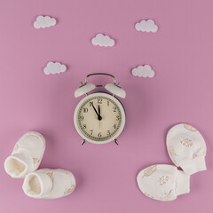 White alarm clock, booties and mittens for a newborn in the clouds on a lilac background. It's five minutes to twelve. It's time to give birth, it's time to go to the maternity hospital. It was flat.