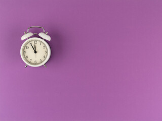 The white alarm clock shows five minutes to twelve on a purple paper background with a copy space. Flat lay. Top view. Place for text. Time: 11.55 am or 23.55 pm