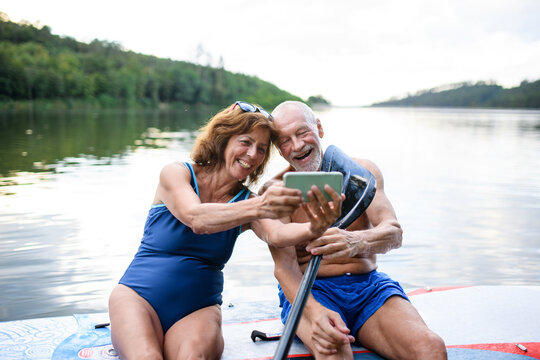 Senior couple sitting on paddleboard on lake in summer, taking selfie with smartphone.