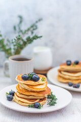 Pancakes laid out in a stack on a plate with blueberries and decorated with greens on a light background. In the background, a slight blur of a milkman, a bouquet of greens with coffee. 