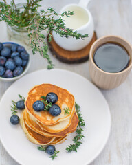 Pancakes laid out in a stack on a plate with blueberries and decorated with herbs on a light background with a milkman, a bouquet of herbs with coffee and berries. Vertical arrangement top view