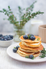Pancakes laid out in a stack on a plate with blueberries and decorated with herbs on a light background with a milkman, a bouquet of herbs with coffee and berries. Vertical arrangement Concept menu