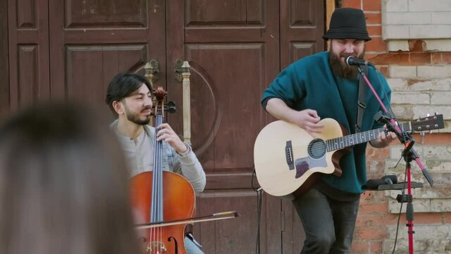 Medium shot of street musicians playing cello and acoustic guitar while performing song for crowd outside