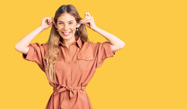 Beautiful caucasian woman with blonde hair wearing summer jumpsuit smiling pulling ears with fingers, funny gesture. audition problem