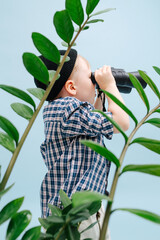 Focused little blond boy looking in binoculars, standing behind plant branches. He's wearing striped long-sleeve shirt and black cap. Studio shot. Over blue background. Side view.