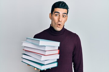 Handsome hispanic man holding a pile of books scared and amazed with open mouth for surprise, disbelief face
